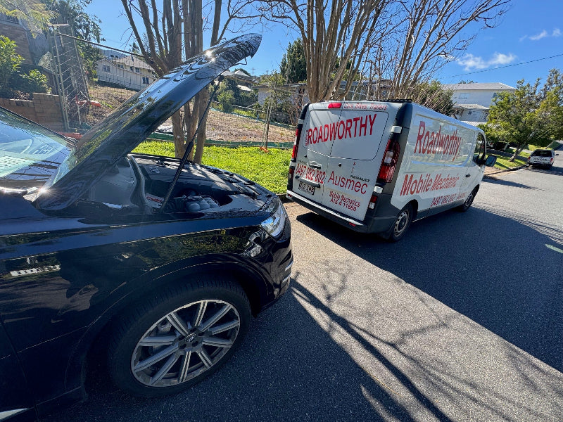 (Car under hood inspection for oil leaks): "Detailed under-hood inspection by Ausmec Mobile Roadworthy ensuring your car meets Carseldine Mobile Roadworthy (Safety Certificate) standards, checking for oil leaks and other vital components." Black car with open hood on a street, next to a white van labeled 'Roadworthy'.