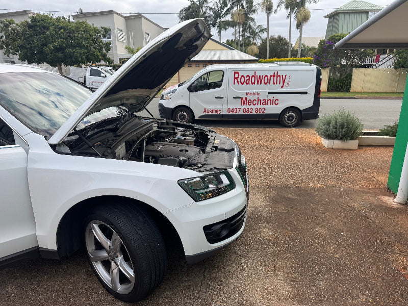 Close-up of an Audi receiving an under-hood inspection for leaks, part of a comprehensive Cashmere Mobile Roadworthy (Safety Certificate) check in a customer's driveway. White car with open hood in a driveway, Roadworthy van in the background