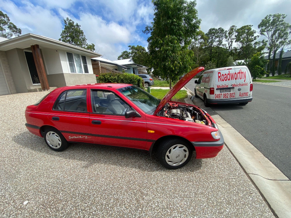A thorough mobile roadworthy inspection of a car in an Eagle Farm customer's driveway, simplifying the process of obtaining a safety certificate. Red car with open hood on a residential street, Roadworthy van in the background.