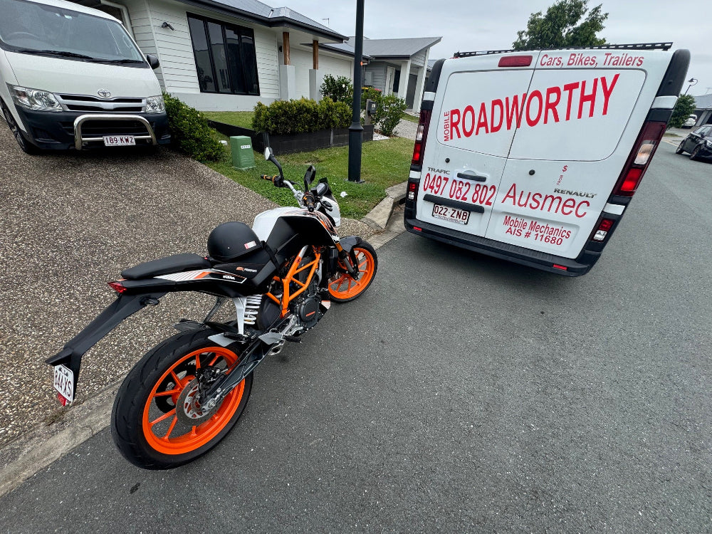An expert performing a mobile roadworthy inspection on a motorcycle at a customer's location in Eagle Farm for their safety certificate. Motorcycle parked on a street with a 'Roadworthy' van in the background.