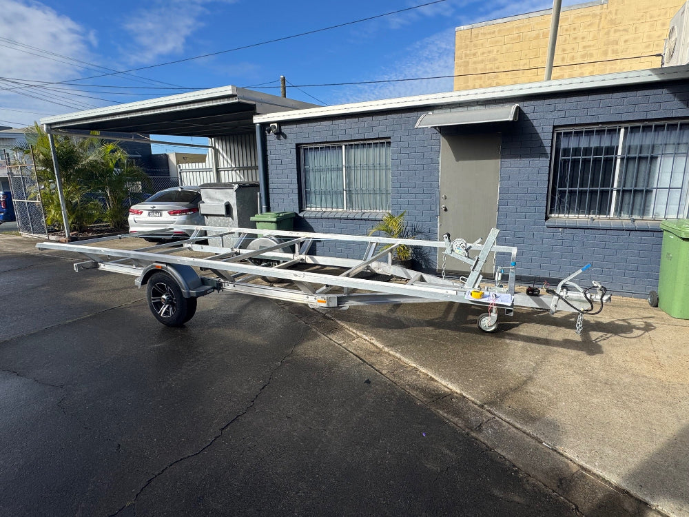Detailed mobile safety certificate inspection of a boat trailer being performed at a customer's home in Everton Hills, ensuring it's fully compliant and roadworthy. Metal boat trailer parked on a concrete driveway next to a building.