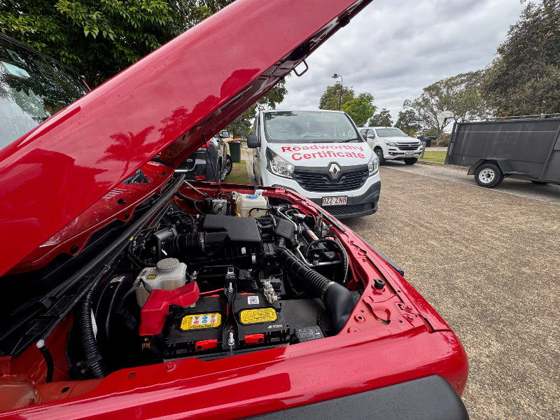 Thorough mobile roadworthy inspection of a car in an Everton Hills customer's driveway, making the process simple for your safety certificate. Open hood of a red vehicle with a van and trailer in the background.