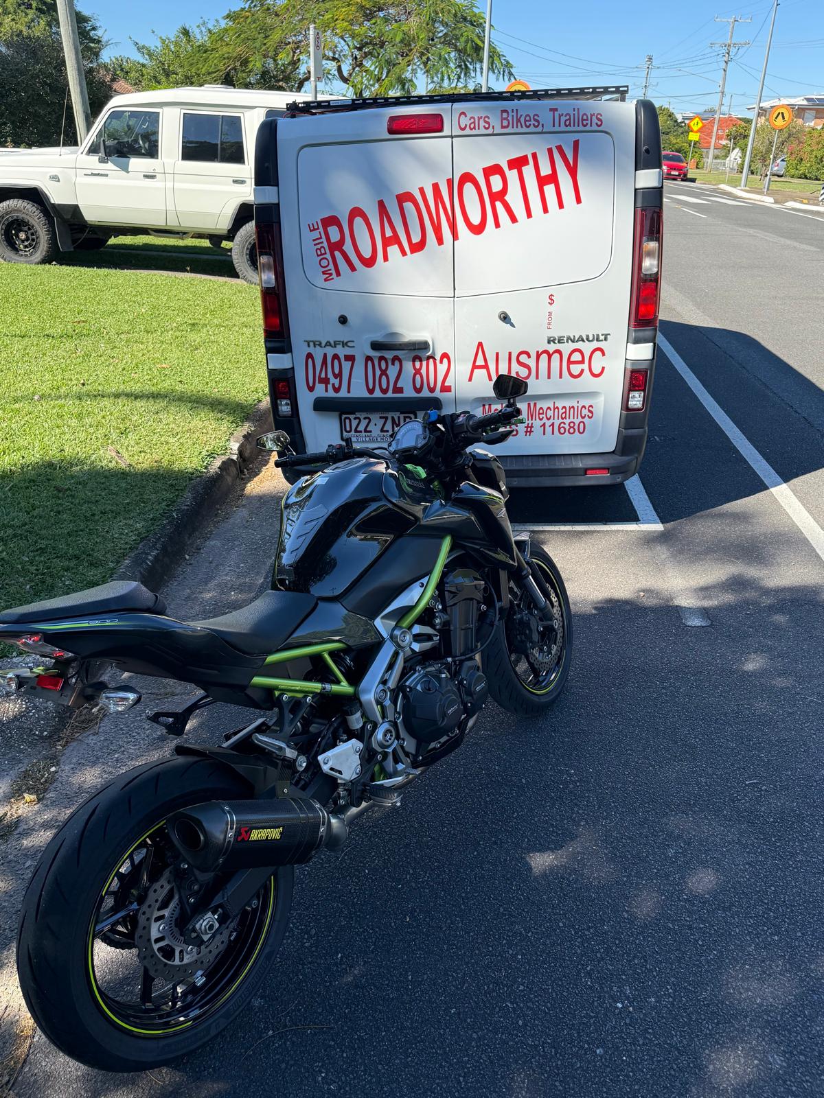Expert conducting a motorcycle mobile roadworthy inspection at a customer's location in Ferny Hills for their safety certificate. Motorcycle parked on a road with a van labeled 'Roadworthy' in the background.