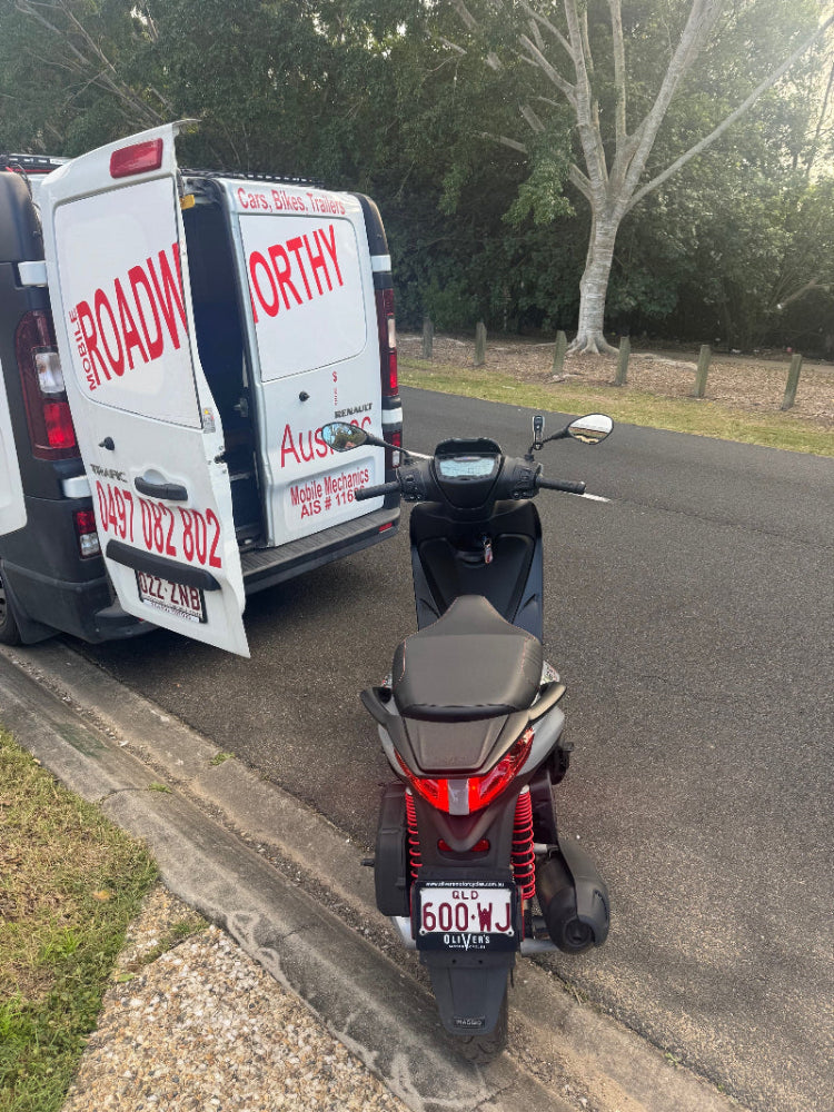 A professional conducting a motorcycle safety certificate inspection on-site in Hamilton, as part of our convenient mobile roadworthy service. A scooter on a road with a van labeled 'ROADWORTHY' in the background.