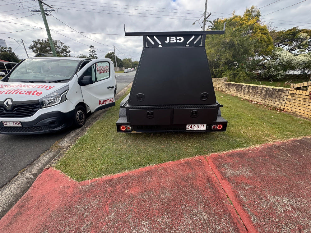 Detailed mobile roadworthy inspection of a box trailer being performed at a customer's home in Hamilton, ensuring a valid safety certificate. Black trailer with 'JBD' branding parked on a grassy area next to a white Roadworthy van.
