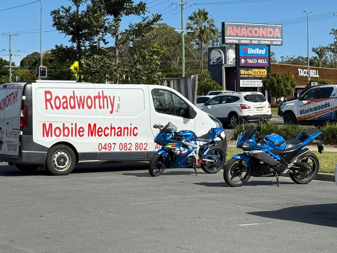 A professional conducting a motorcycle mobile roadworthy inspection at a customer's location in Hendra for their safety certificate. Roadworthy Mobile Mechanic van with two motorcycles on a street.