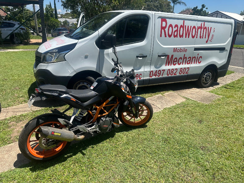 "Motorcycle RWC being performed right next to our Ausmec mobile van for a fast Redcliffe Mobile Roadworthy (Safety Certificate)." Motorcycle parked on grass with a 'Roadworthy' van in the background