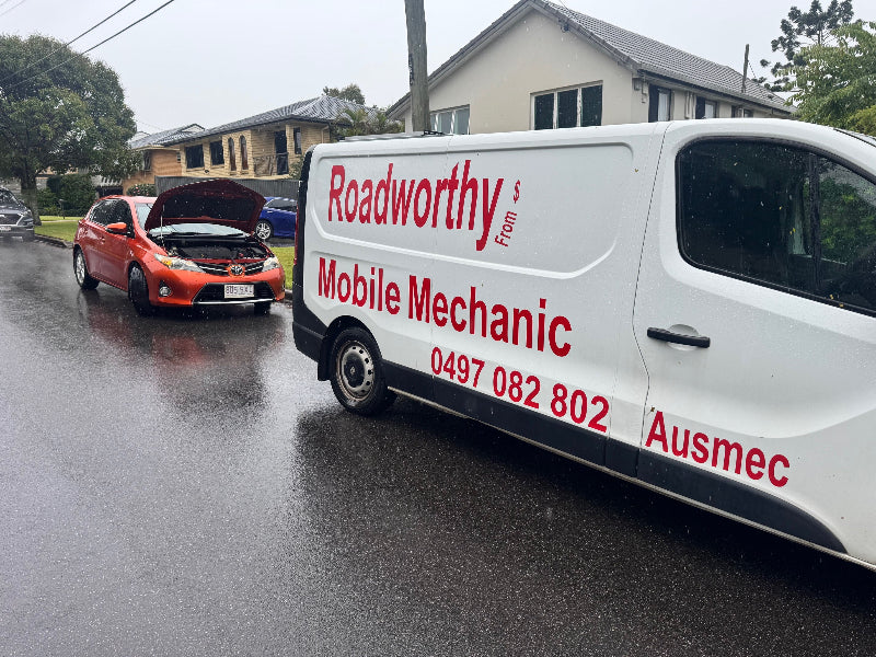 "Toyota car receiving a Narangba Mobile Roadworthy (Safety Certificate) inspection on a rainy day, demonstrating our all-weather service." White van with 'Roadworthy Mobile Mechanic' branding on a rainy street.