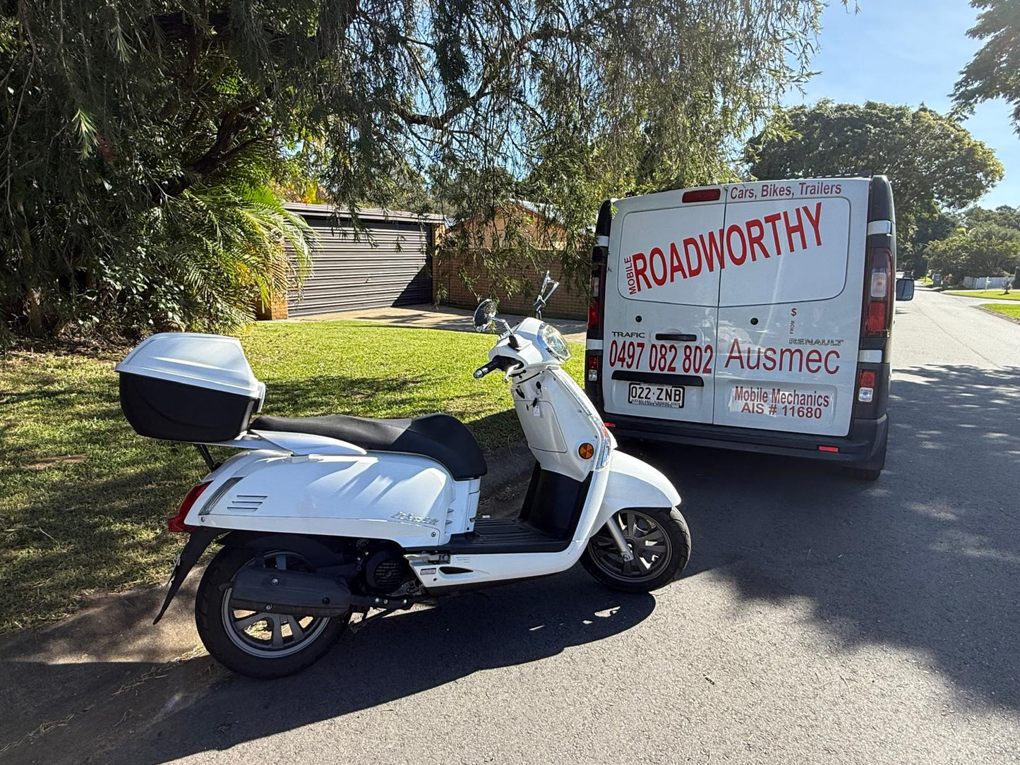 An expert conducting a detailed mobile roadworthy inspection on a motorcycle at a customer's location in Joyner for their safety certificate. White scooter parked on a road with a van labeled 'Roadworthy Ausmec' in the background.