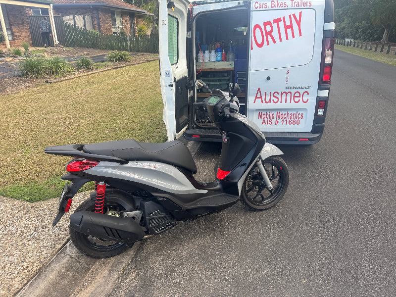 A moped motorcycle visible behind the Ausmec Mobile Roadworthy van, ready for a Kurwongbah mobile roadworthy (safety certificate) inspection. Scooter parked on a driveway with a van labeled 'Ausmec' in the background.
