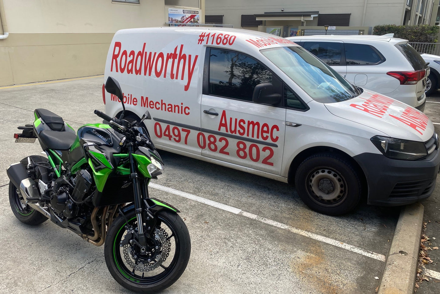 "Motorcycle undergoing a thorough Burpengary Mobile Roadworthy (Safety Certificate) inspection in a parking bay." White van with red text and a motorcycle on a concrete surface
