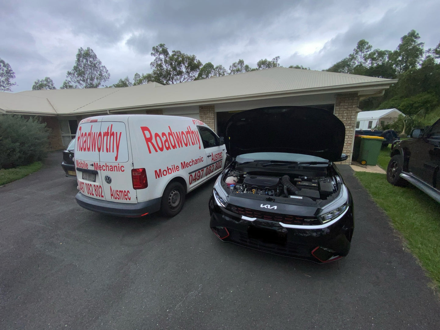 An Ausmec Mobile Roadworthy inspector performing a comprehensive safety certificate inspection on a car in a customer's driveway, highlighting our convenient Newport mobile roadworthy service. A vehicle with an open bonnet receiving a roadworthy inspection next to a van with the Ausmec Mobile RWC & Mechanics branding.