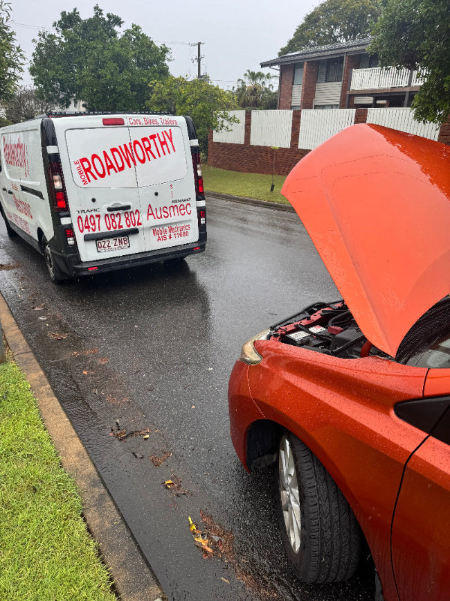 "Dedicated mechanic performing a car inspection for a Sandgate Mobile Roadworthy (Safety Certificate), even in cold and rainy weather." Orange car with a lifted hood on a residential street, with a van labeled 'Roadworthy' in the background.