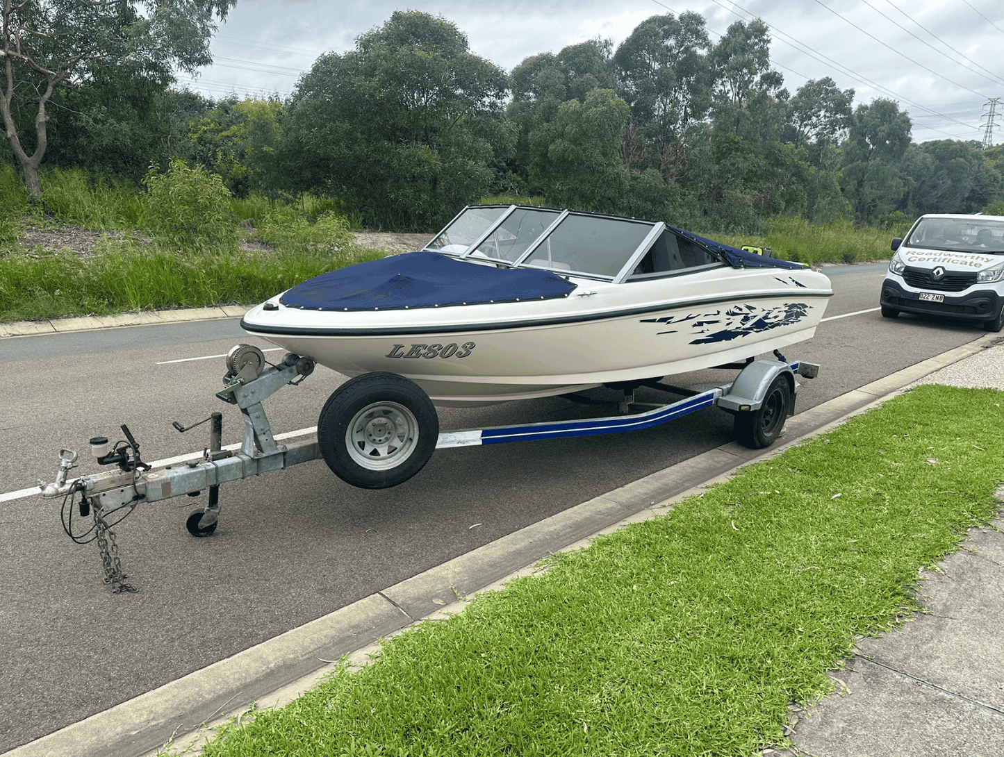 "Boat trailer ready for its Burpengary East Mobile Roadworthy (Safety Certificate) inspection at a customer's Burpengary East property." White boat with blue cover on a trailer by a road with greenery in the background