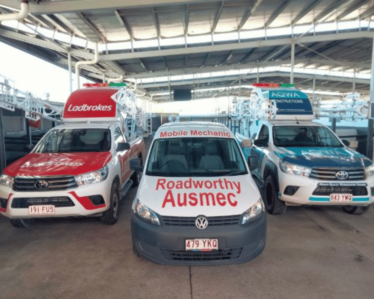 "Ausmec Mobile Inspection van ready to provide Caboolture South Mobile Roadworthy (Safety certificate) service for multiple vehicles." Three vans, two with 'Roadworthy Ausmec' branding and one with 'Lodbrokes' branding, parked under a large industrial or warehouse roof.
