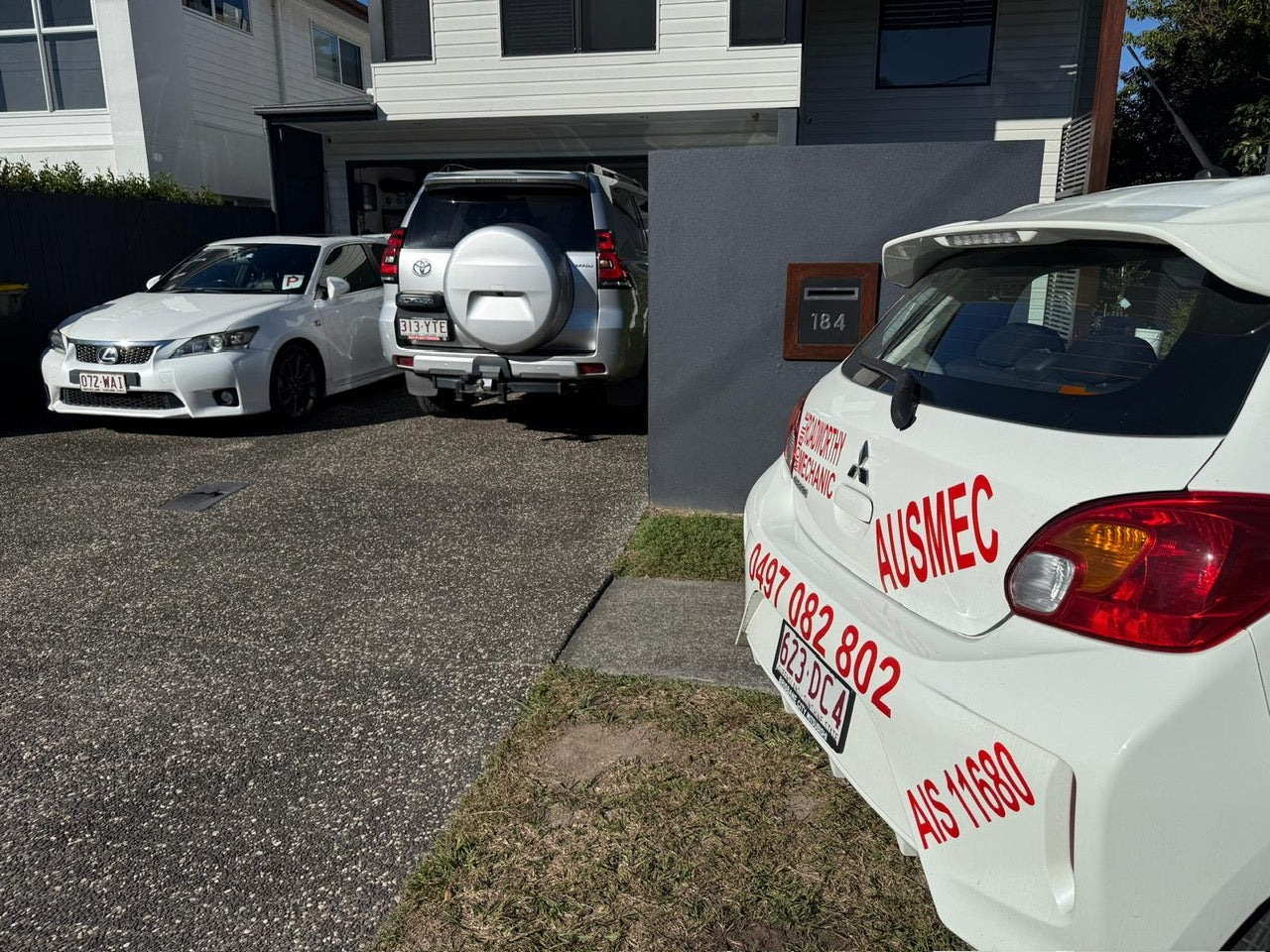"Two cars in Bellmere undergoing Bellmere Mobile Roadworthy (Safety Certificate) inspections in a customer's driveway for interstate registration transfer." Two cars parked in a driveway with a close-up of a white car's rear end showing 'AUSMEC' branding.