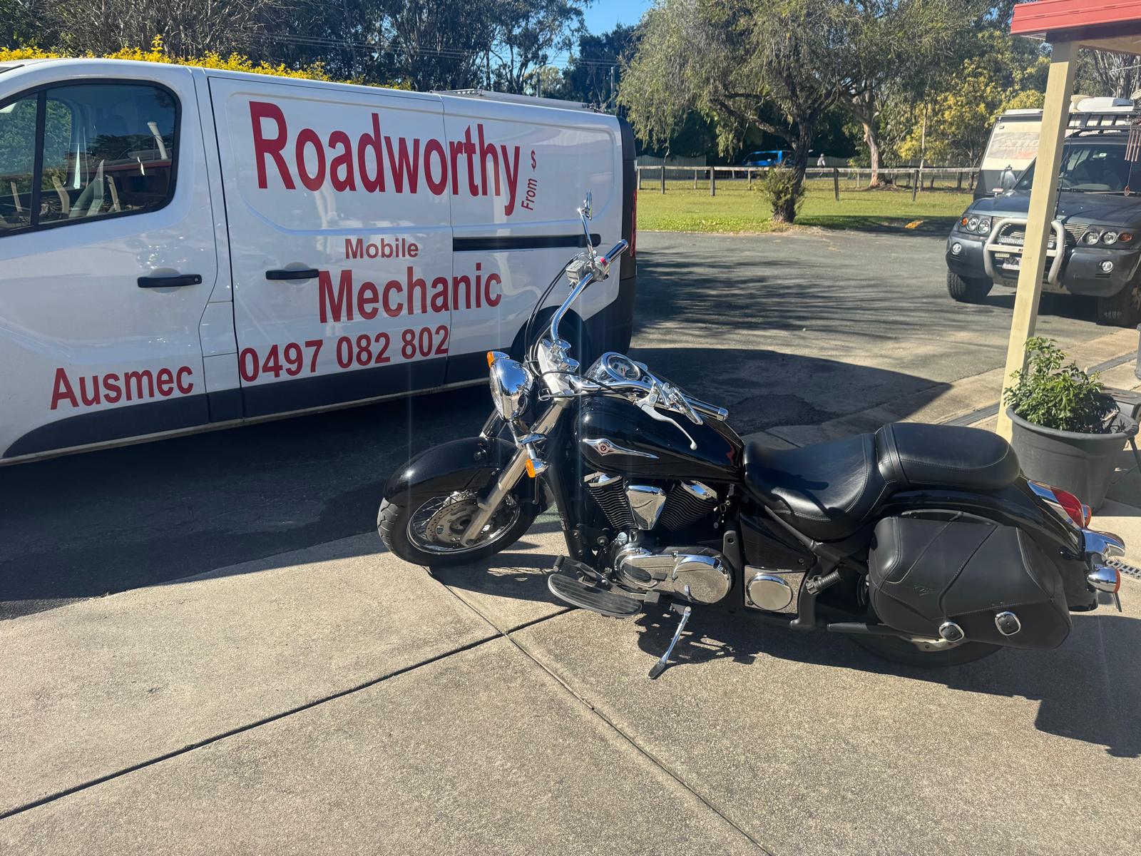 A close-up view of an Ausmec Mobile Roadworthy inspector meticulously examining a motorcycle during an Ascot Mobile Roadworthy (Safety Certificate) assessment. Black motorcycle parked next to a Roadworthy van in a parking lot.