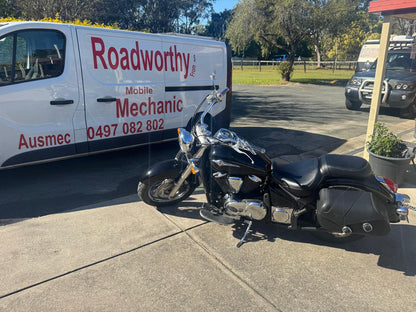 A close-up view of an Ausmec Mobile Roadworthy inspector meticulously examining a motorcycle during an Ascot Mobile Roadworthy (Safety Certificate) assessment. Black motorcycle parked next to a Roadworthy van in a parking lot.