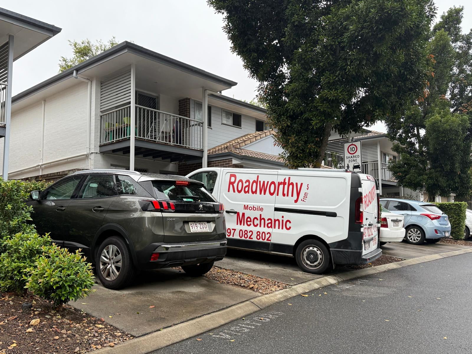 (Peugeot being inspected at townhouse complex): An Ausmec Mobile Roadworthy inspector meticulously examining the front wheel and tire of a silver Peugeot sedan parked in front of a modern townhouse complex, demonstrating convenient on-site Banyo Mobile Roadworthy (Safety Certificate).