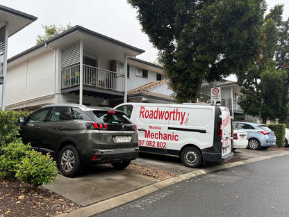 (Peugeot being inspected at townhouse complex): An Ausmec Mobile Roadworthy inspector meticulously examining the front wheel and tire of a silver Peugeot sedan parked in front of a modern townhouse complex, demonstrating convenient on-site Banyo Mobile Roadworthy (Safety Certificate).