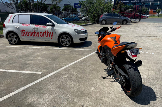 Orange motorcycle parked in a parking lot with a white car labeled 'Roadworthy' in the background in Brendale.