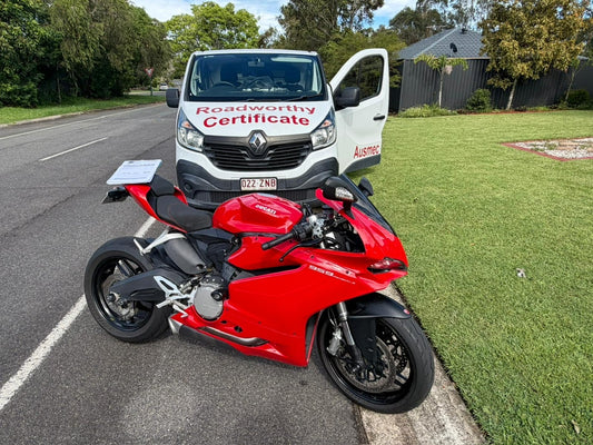 Red motorcycle parked on a road with a van labeled 'Roadworthy Certificate' in the background in Clayfield