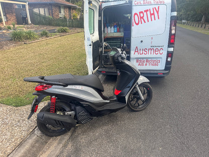 A motorcycle or moped is undergoing a thorough North Lakes Mobile Roadworthy (Safety Certificate) inspection. Our easily identifiable mobile van is parked nearby, showcasing our on-site service capability."