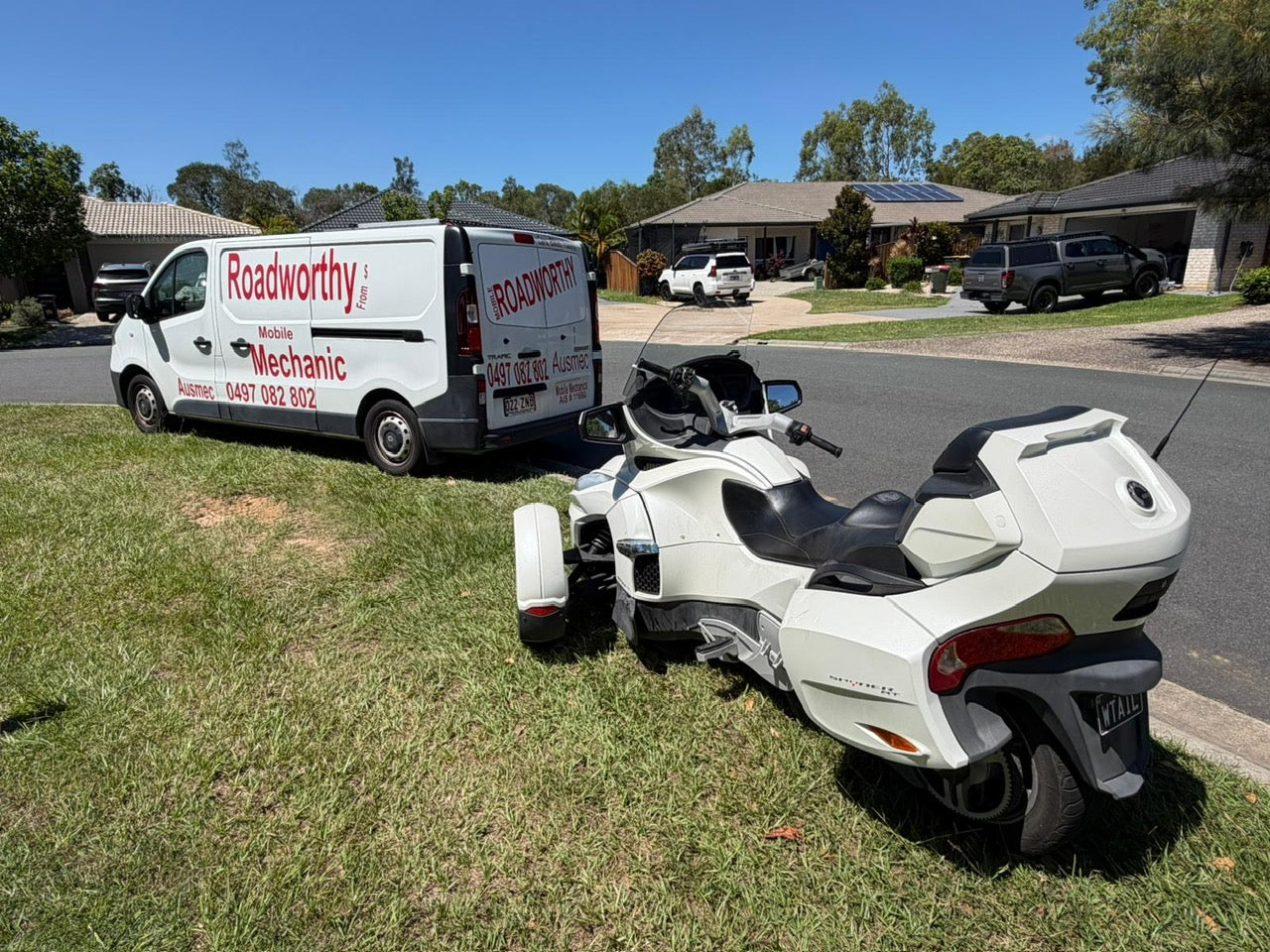 White motorcycle on grass with a van labeled 'Roadworthy' in the background in Enoggera