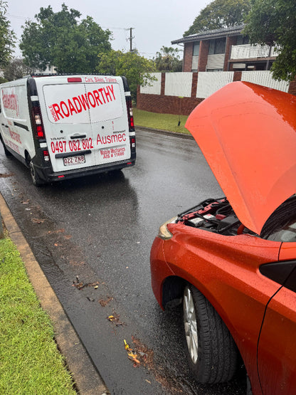 "Dedicated mechanic performing a car inspection for a Sandgate Mobile Roadworthy (Safety Certificate), even in cold and rainy weather." Orange car with a lifted hood on a residential street, with a van labeled 'Roadworthy' in the background.