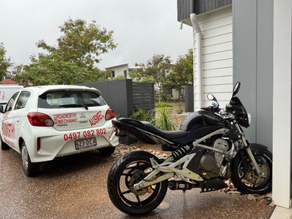 White car with 'Ausmec Mobile Roadworthy & Mechanics' branding parked next to a black motorcycle on a wet driveway in Sandgate.