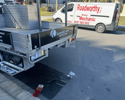 "Ute defects being repaired at a customer's home as part of the comprehensive Morayfield Mobile Roadworthy (Safety certificate) service."
Rear of a Ute with 'LandCrate' branding and a white van with 'Roadworthy' branding in the background.